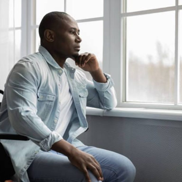 Lonely disabled person at home or rehabilitation clinic during COVID-19 lockdown. Melancholic adult african american man in wheelchair looking at window and dreaming, in living room interior, profile