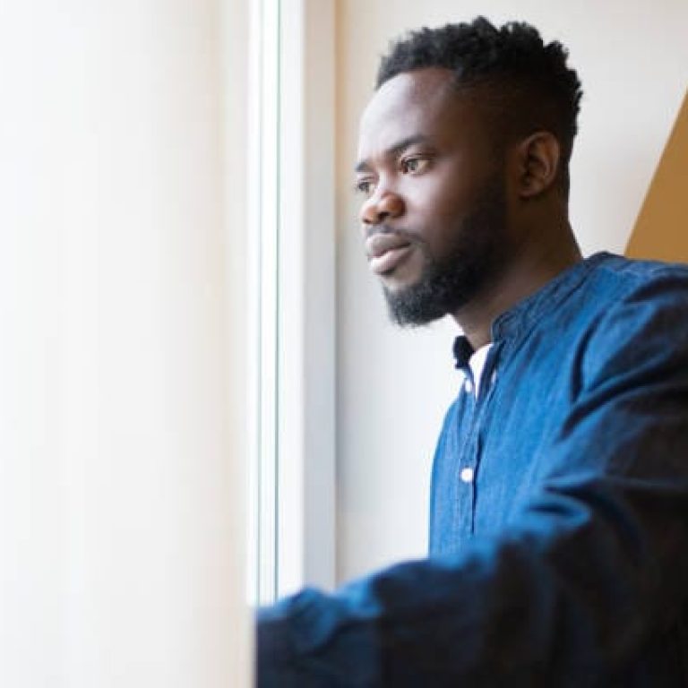 A worried young man is looking thoughtful through the bright window.