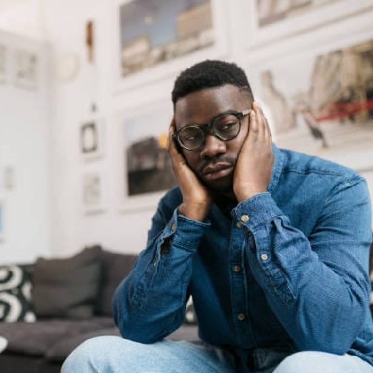Sad and lonely, young and depressed African American man sitting at alone home, worrying about something and resting his head between the palms of his hands