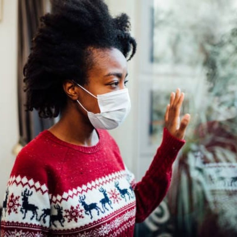 An African American woman wearing a protective face mask, looking melancholic, is looking trough the window of her home, wearing a protective face mask