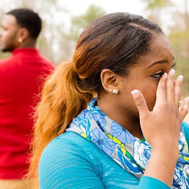 Emotions. Young African descent woman and man stand with backs to each other after an arguement. They are distraught after a fight.  She is crying from despair and is sad and upset.   Man looks away with arms crossed in background.  Outdoors in winter season.  Head and shoulders.