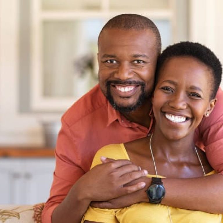 Happy mature black couple bonding to each other and smiling while sitting on couch. Portrait of smiling black man embrace his wife from behind and looking at camera. Smiling husband and beautiful woman laughing.