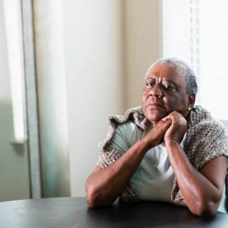 A lonely senior African-American woman in her 70s sitting at home at a table by a window. She is looking at the camera with a serious expression, elbows on the table and hands clasped.