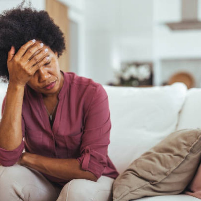 A middle-aged woman sitting on a white sofa, appearing stressed and worried, is holding her head with one hand. The setting is a warm, cozy living room environment.