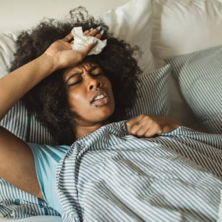 Young African-American woman feeling sick at home. Lying in bed, drinking tea and medicine. Daytime.